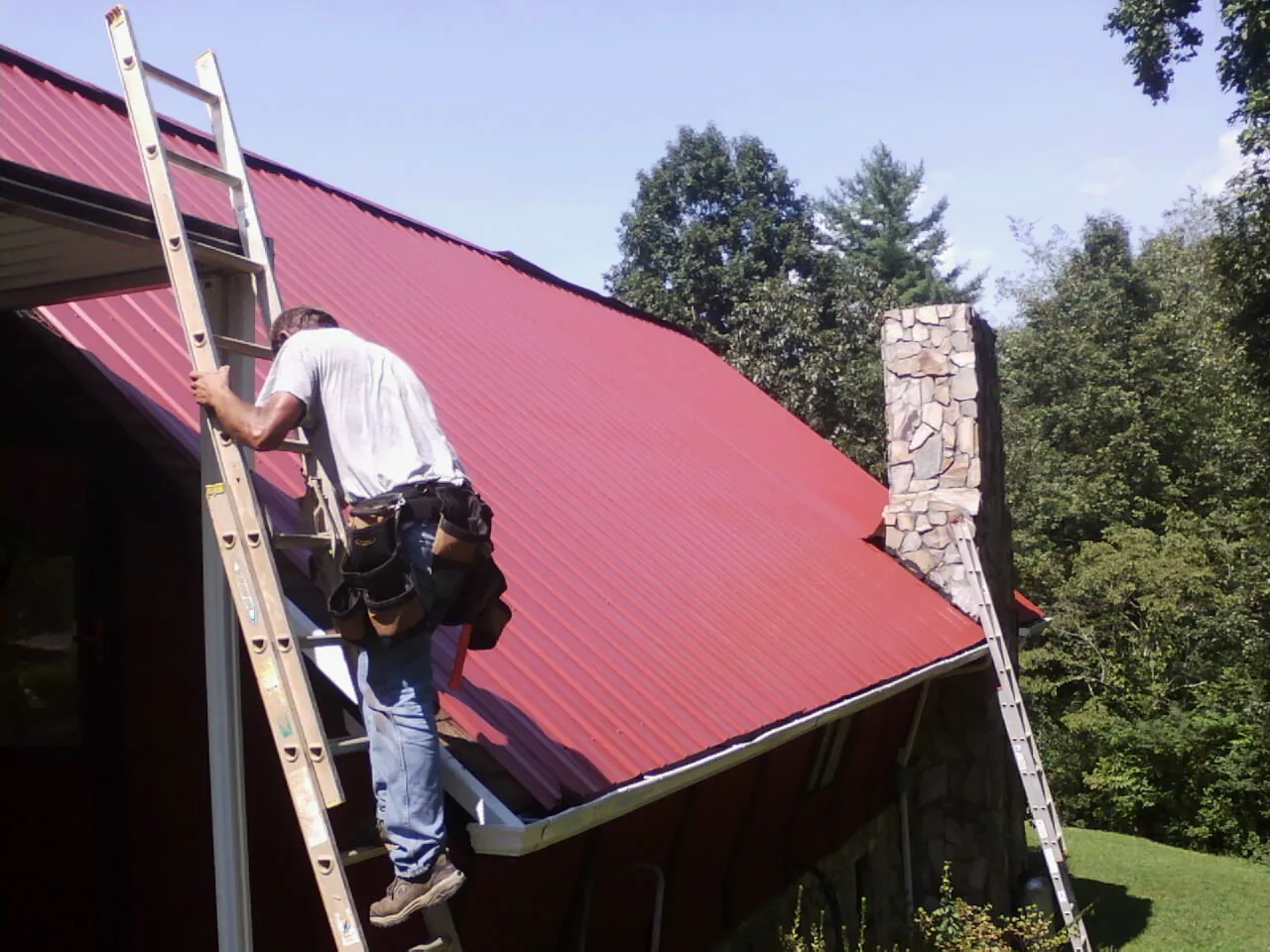 Roofer on a ladder beside a red metal roof and stone chimney — Soffit & Fascia Repair by Roofing Brothers in Mercer